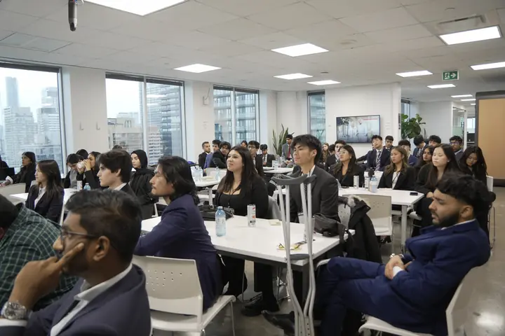 Competitors seated at long white tables in a bright office, city towers through the windows.