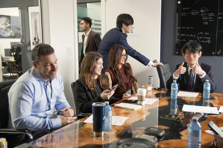 Five people around a glass table with water bottles and papers; one gives a thumbs up.